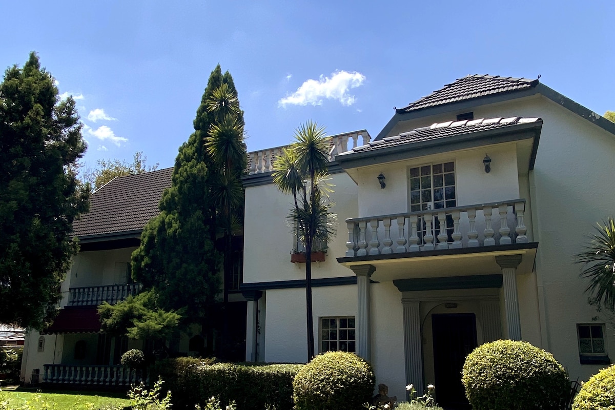 The exterior of a charming two-story house is showcased, featuring white walls and a sloping roof. Lush greenery, including tall trees and manicured bushes, surrounds the property. A balcony with decorative railings adds character to the facade, complemented by clear blue skies in the background.