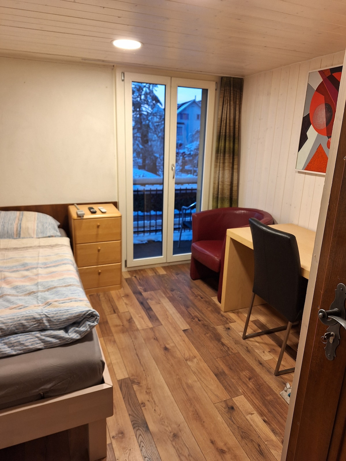 A cozy room features a spacious bed with light bedding, a nightstand, and a chest of drawers. A workspace with a desk and chair is positioned across from the bed. Natural light enters through double glass doors that lead to a small balcony.