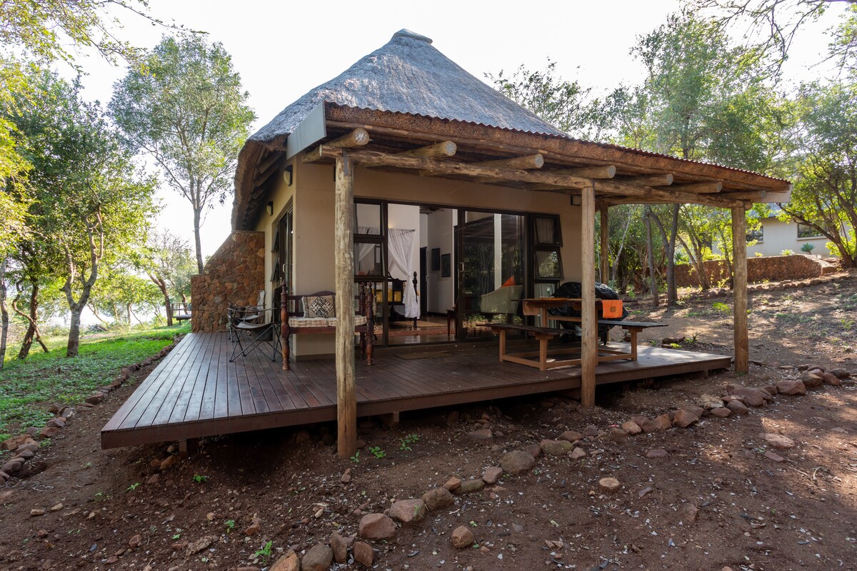 The studio's exterior is characterized by a thatched roof and wooden deck, blending harmoniously with the surrounding greenery. A covered patio provides an inviting space for outdoor seating, while large windows offer views of the nearby ravine and natural landscape.