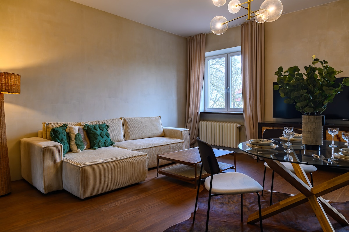 The spacious living area features a comfortable sofa adorned with decorative pillows, paired with a stylish dining table set for four. Natural light enters through the window, complementing the neutral wall tones and modern lighting fixture. A TV and decorative plants add to the inviting decor.