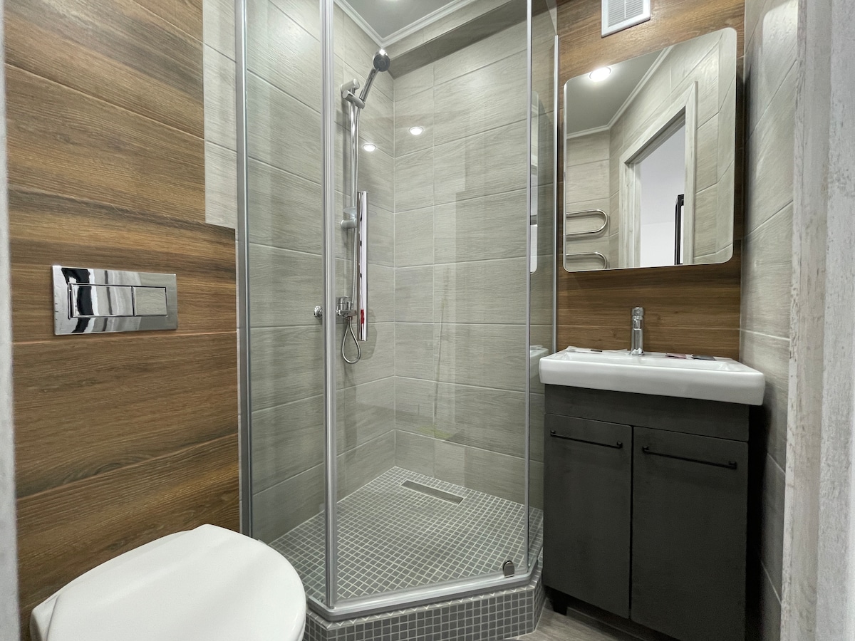 A modern bathroom is presented with a glass-enclosed shower featuring sleek fixtures. Earth-toned tiles are complemented by a dark vanity with a sink and mirror. A white toilet is located beside the shower, and natural light is softly filtered through the frosted glass.