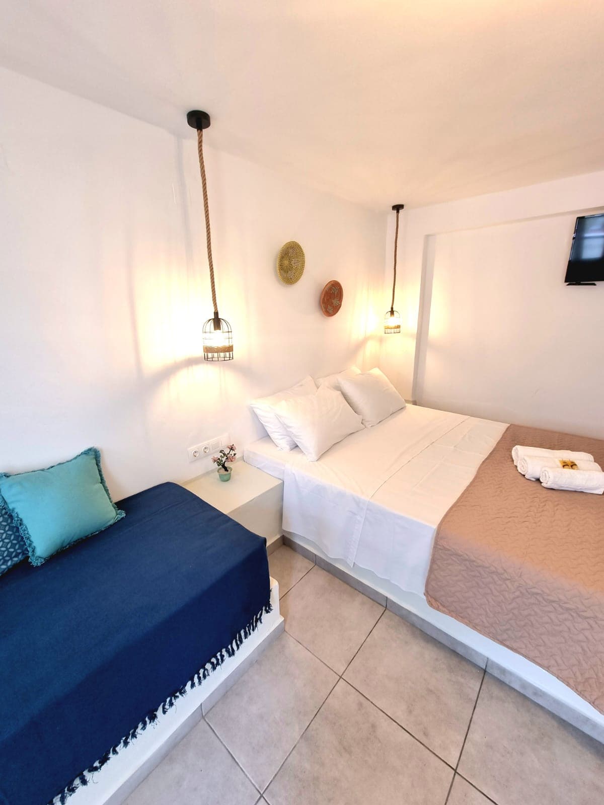 A bright sleeping area features two beds: one spacious bed dressed in white linens and a second smaller bed adorned with a blue throw. Decorative wall hangings and pendant lights add subtle touches to the room, complemented by natural light streaming in.