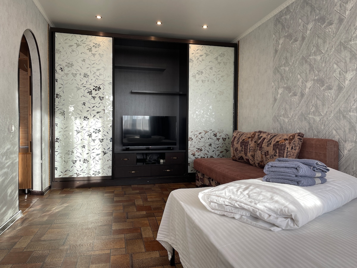 A spacious room features a sleek entertainment unit with a flat-screen TV, flanked by decorative panels. A comfortable sofa is positioned nearby, complemented by neatly stacked towels on a cozy bed. Natural light floods the room through large windows, enhancing the inviting atmosphere.