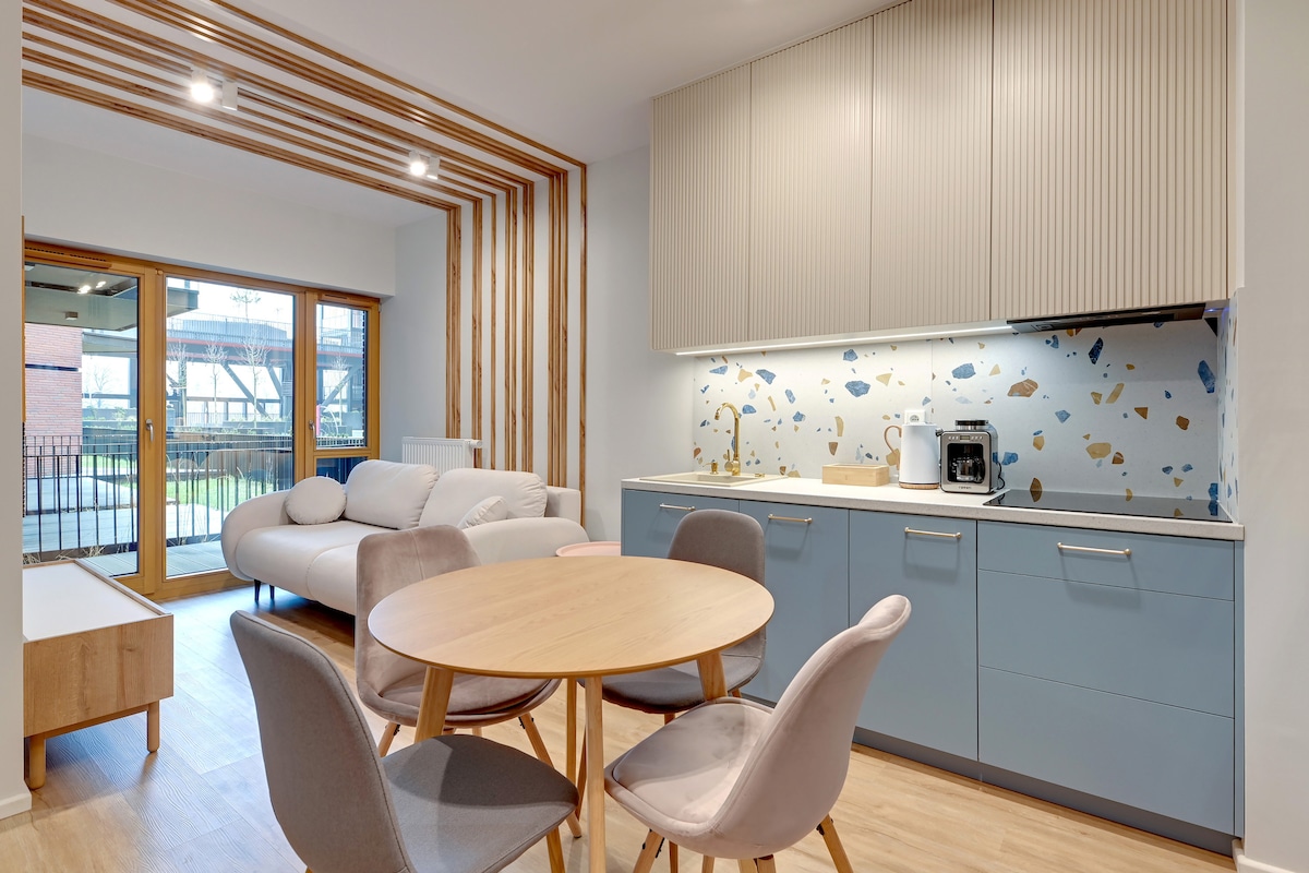 An open-plan living space is shown, featuring a modern kitchen area with light blue cabinetry and decorative wall tiles. A round dining table with four chairs occupies the center, while a comfortable sofa is positioned nearby. Large windows allow natural light to fill the room.
