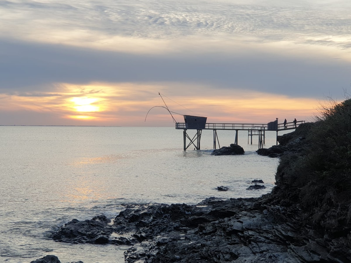 A fishing pier extends over calm waters, silhouetted against a sunset. Soft hues of orange and pink reflect on the surface, while the rocky shoreline frames the foreground. Two figures are visible on the pier, enjoying the tranquil evening ambiance.