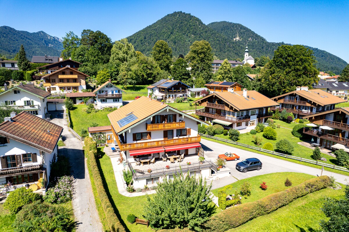 The image showcases a freestanding vacation house situated in a tranquil setting, surrounded by lush greenery and mountains. A spacious terrace is visible in the front, with several balconies accessible from the upper levels, offering ample outdoor space in a serene neighborhood.