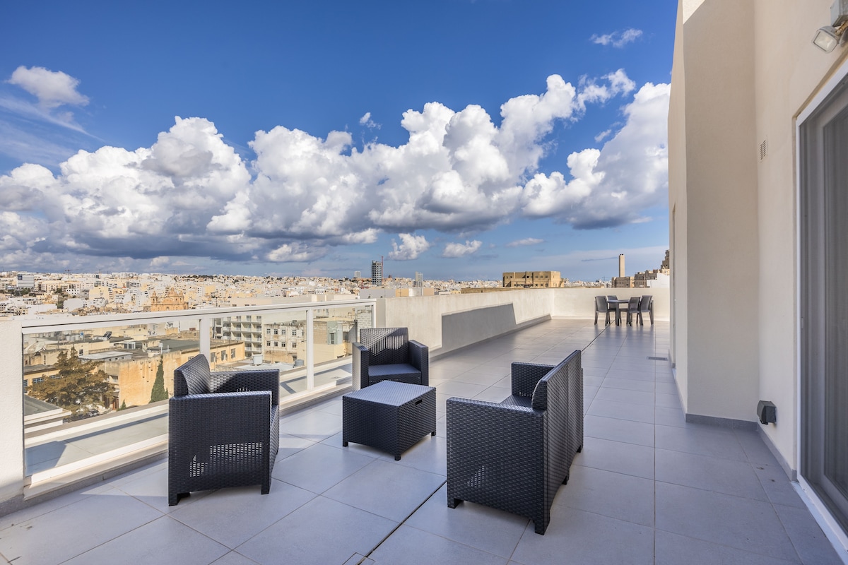 A spacious outdoor terrace is furnished with comfortable seating arrangements and a coffee table, offering panoramic views of the city and sky. The floor is tiled, enhancing the modern aesthetic, while the surrounding balustrade ensures safety and unobstructed sightlines to the vibrant landscape.