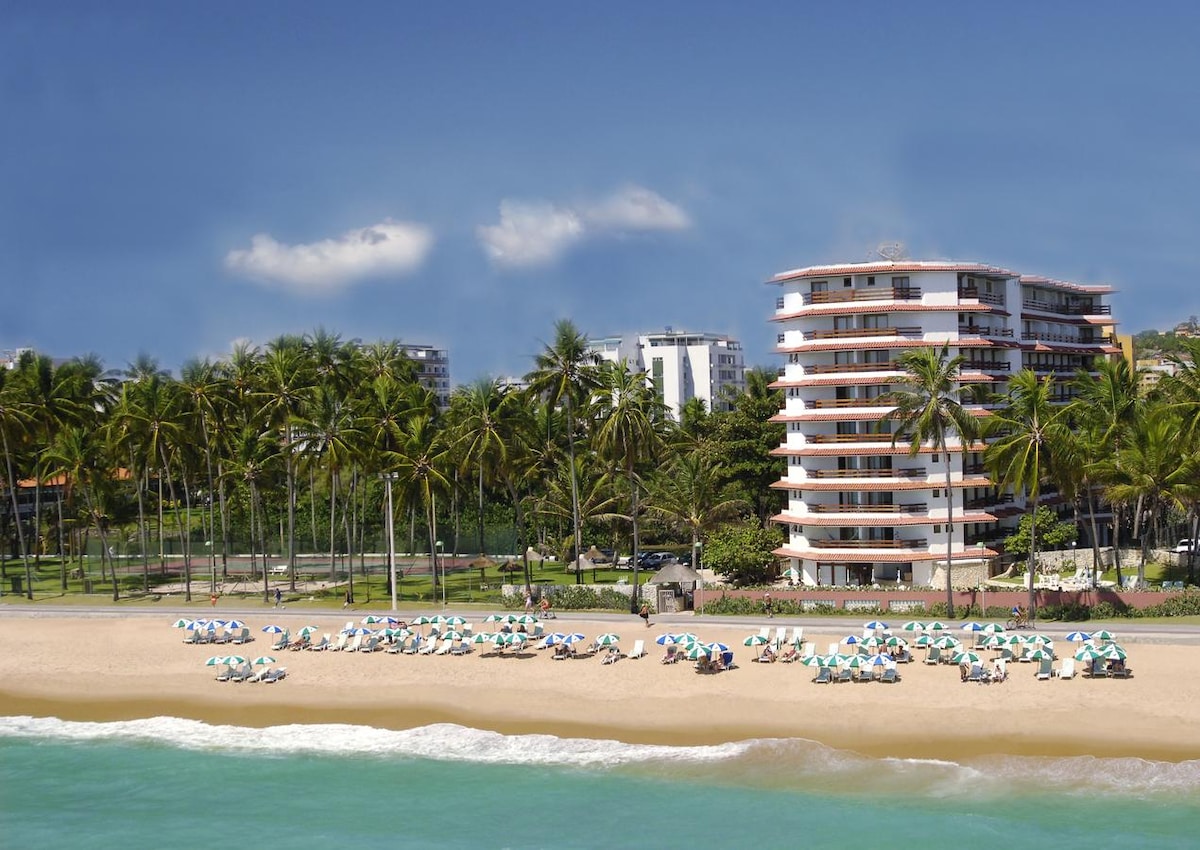 The beachfront resort is featured with loungers arranged under umbrellas on sandy shores. Palm trees line the beach, providing shade. The modern building stands in the background, overlooking the ocean, with clear skies and a hint of distant cityscape visible beyond the greenery.