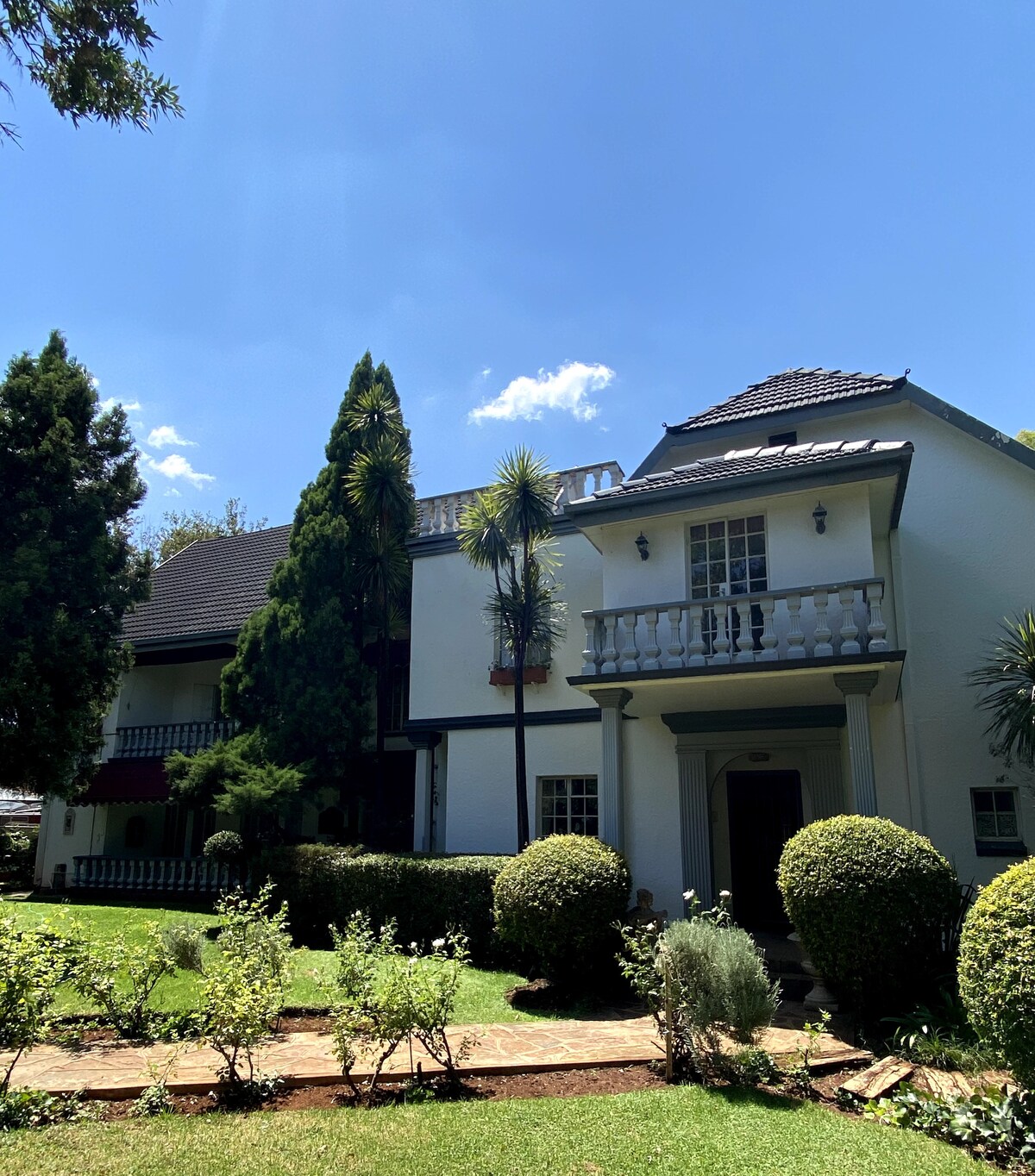 The exterior of a charming two-story house is showcased, featuring a white facade with dark roofing. Lush gardens surround the property, with neatly trimmed bushes and a stone pathway leading to the entrance. Trees provide shade under a clear blue sky.
