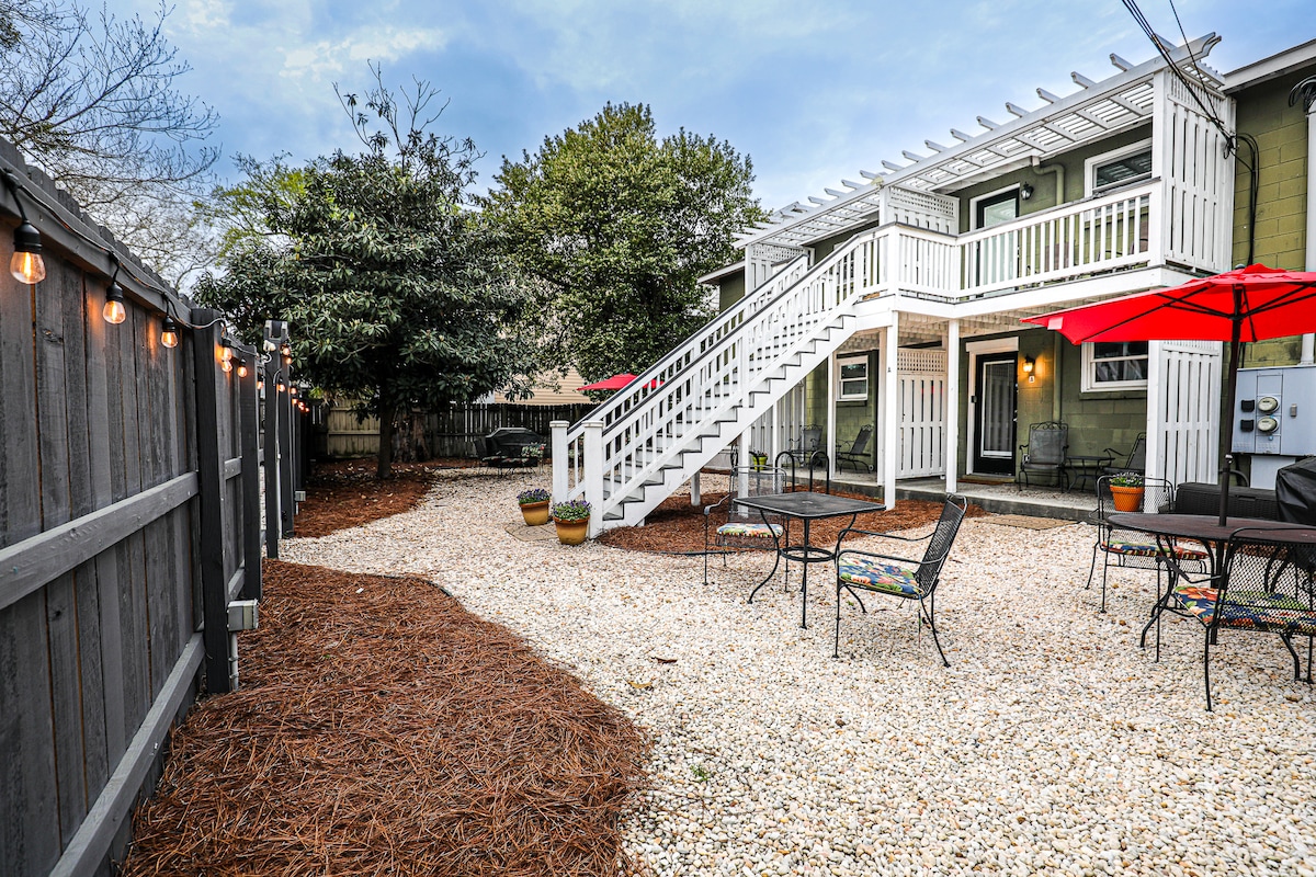 An outdoor space features a gravel area surrounded by greenery and decorative lights. Patio tables with chairs are arranged beneath red umbrellas, providing shaded seating options. Steps lead to the upper unit, offering a structured balance to the inviting environment.