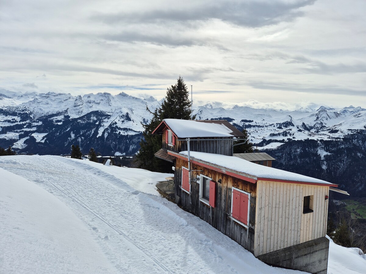 A charming mountain cottage is surrounded by a blanket of snow, positioned against a stunning backdrop of snow-capped peaks. The structure features distinct wooden exteriors with a sloping roof, while the tranquil landscape extends into the distance, highlighting the serene alpine environment.