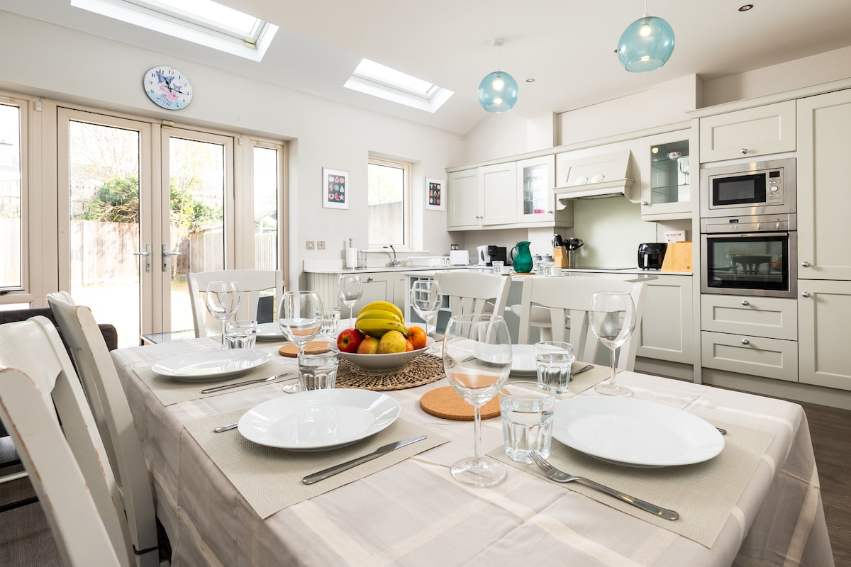 The kitchen features a light-filled dining area with a table set for four, showcasing elegantly arranged glassware and cutlery. A bowl of fresh fruit adds a pop of color, while the modern kitchen includes sleek cabinetry and stainless-steel appliances. Large patio doors lead to the garden.