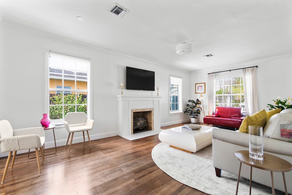 A spacious living room is showcased, featuring a white sofa and a red leather couch. A wall-mounted flat screen TV is positioned above a streamlined fireplace. Natural light is welcomed through two windows, and a plush area rug complements the hardwood flooring.