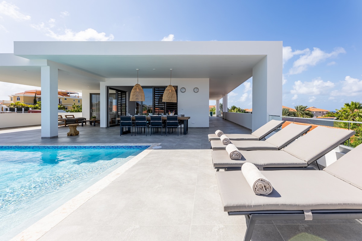 A modern outdoor terrace featuring a sparkling pool alongside spacious lounge chairs. The area is complemented by a dining table under a stylish canopy. Ample natural light and a clear blue sky enhance the inviting setting, showcasing a seamless connection between indoor and outdoor spaces.