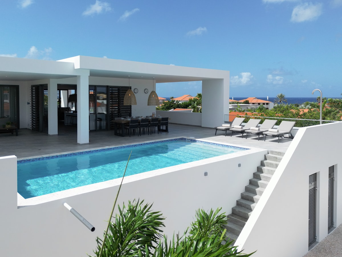 A modern villa exterior features a sparkling pool surrounded by several lounge chairs. A spacious terrace is visible, leading to a dining area under pendant lighting. The ocean can be seen in the background, and steps guide the way from the terrace to the pool area.