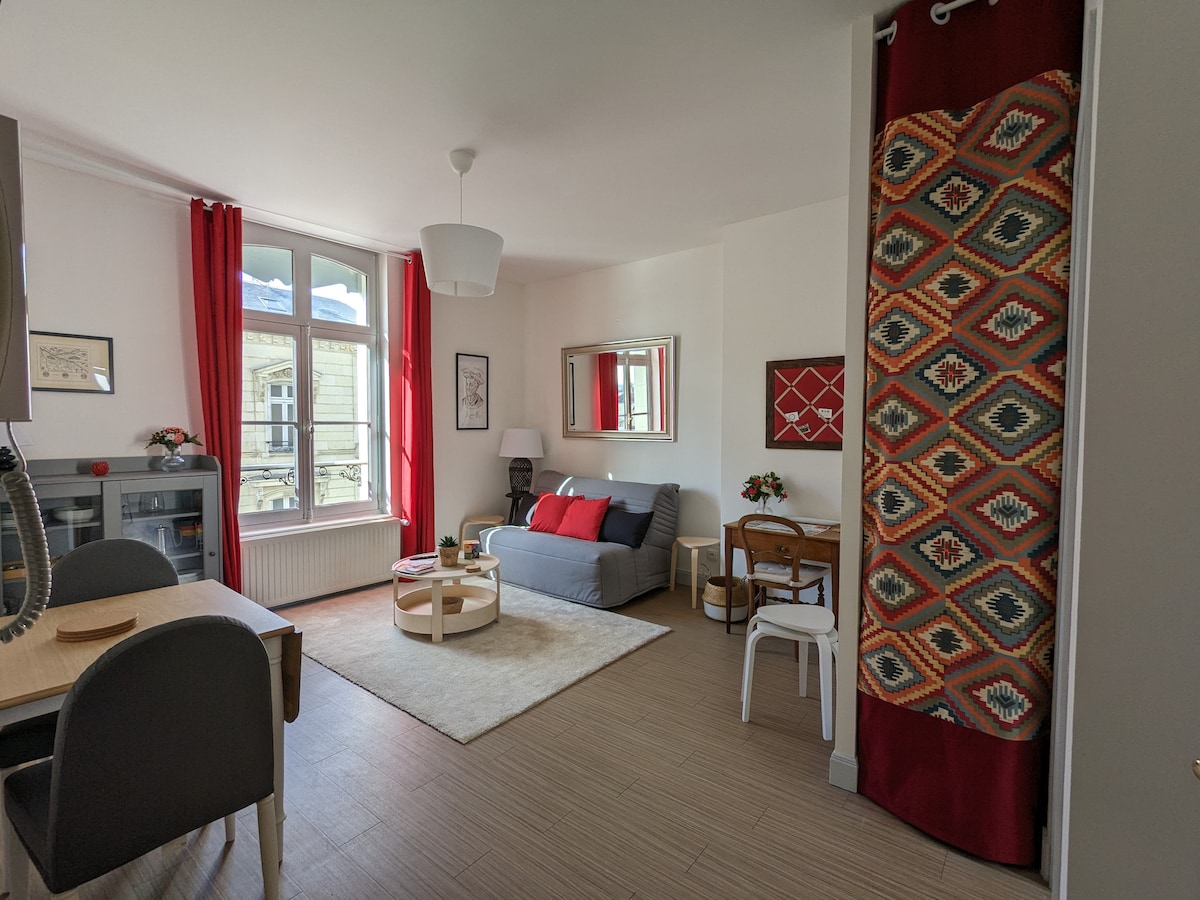 The living area features a spacious sofa with red and gray accents, complemented by a round coffee table. Large windows, dressed in vibrant red curtains, allow natural light to fill the room. A dining table with chairs is visible nearby, creating a cozy gathering space.
