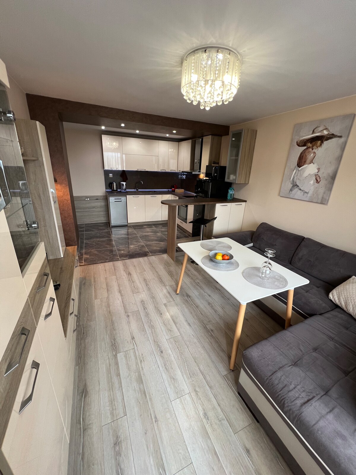 A modern living space is showcased, featuring a gray sectional sofa and a light wooden coffee table with a bowl of fruit. A well-equipped kitchen is visible in the background, with sleek cabinetry and contemporary appliances. Natural light illuminates the area through large windows.