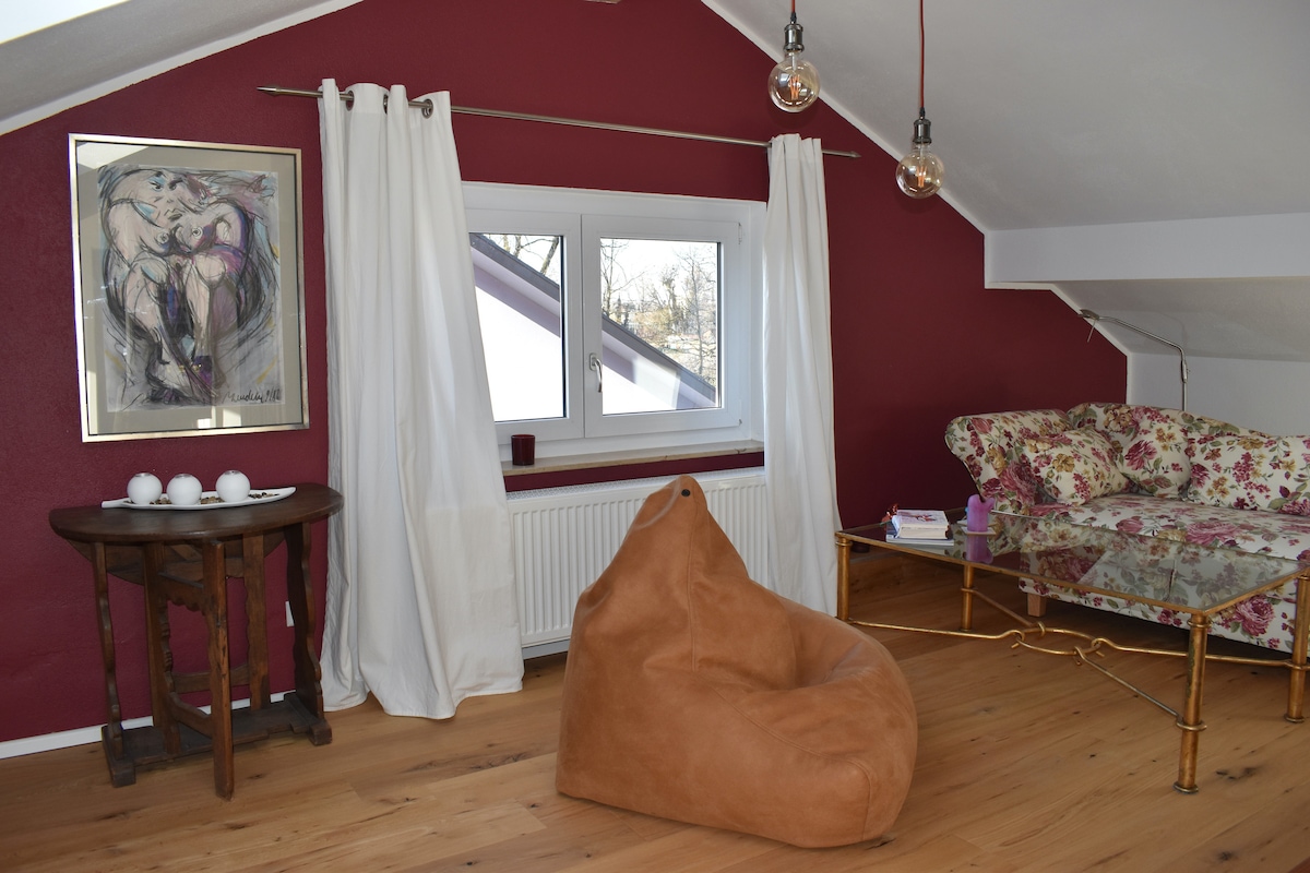 A seating area is highlighted by a floral-patterned couch and a cozy bean bag chair. A small round table holds several white ceramic cups, while large windows framed by white curtains allow natural light to brighten the space. A piece of art adorns the wall.