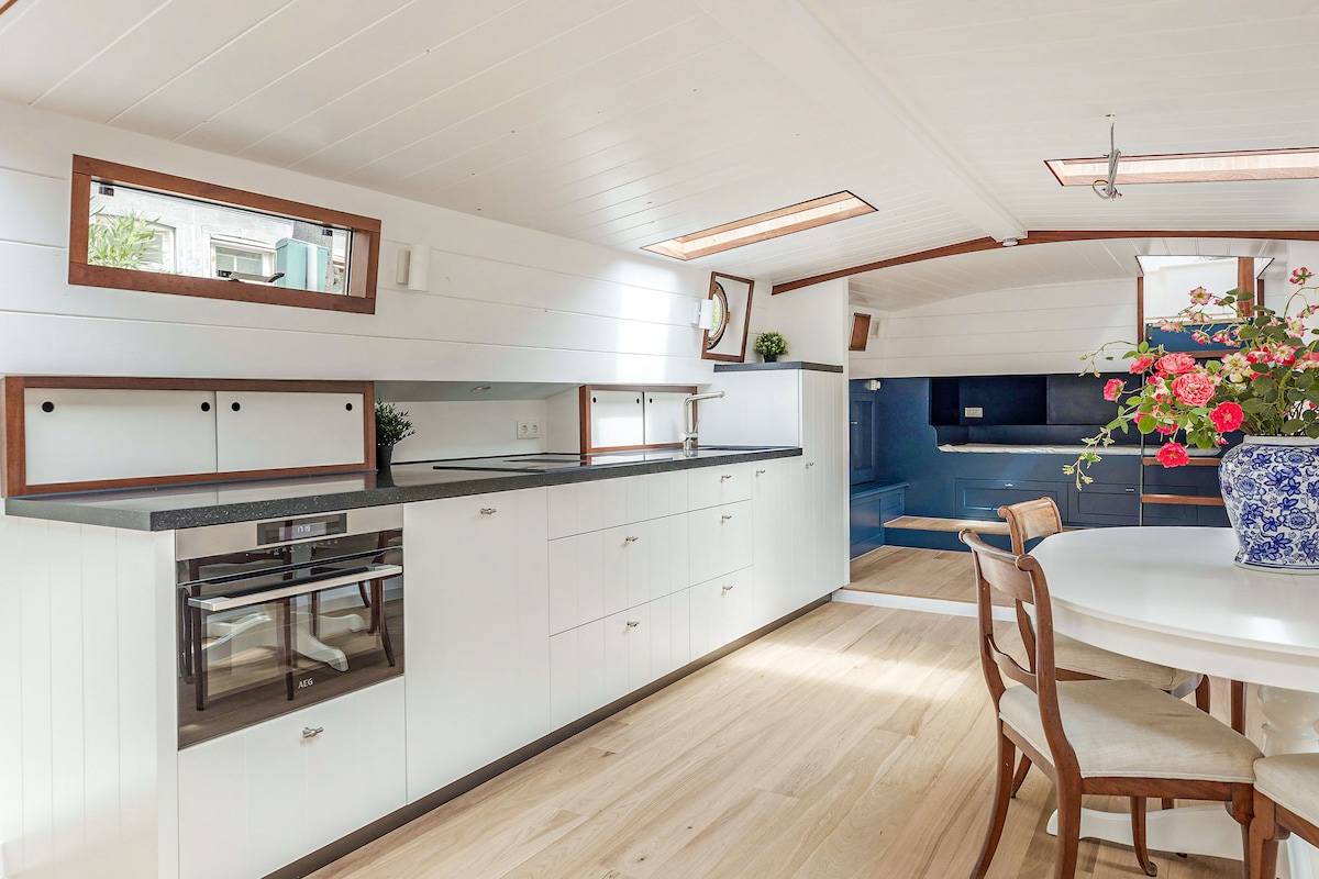 An open kitchen is visible, featuring modern cabinetry and appliances, including an oven and a spacious preparation area. Natural light floods through overhead windows, highlighting the wooden flooring and the adjacent dining area equipped with a round table and wooden chairs.