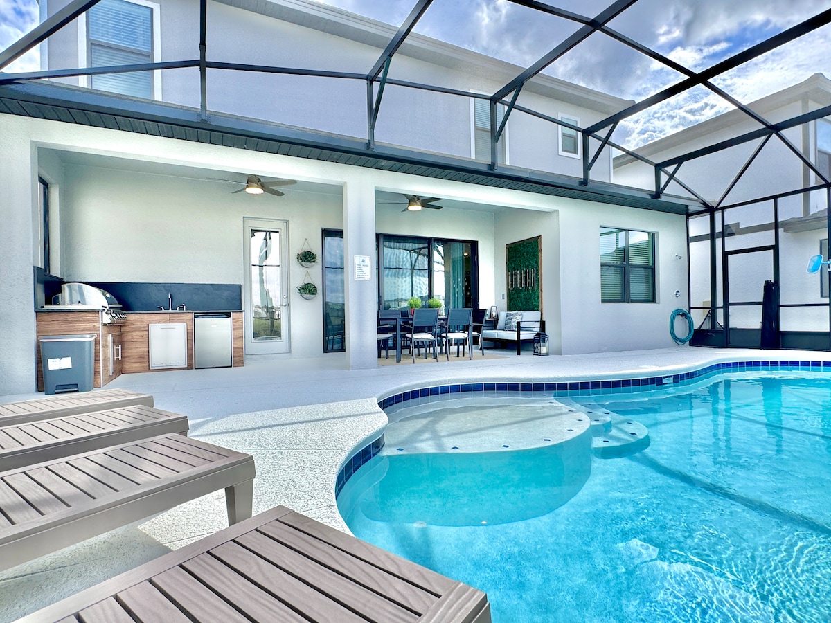 A private leisure area is visible, featuring a sparkling pool with a built-in spa. Comfortable lounge chairs surround the pool, and a summer kitchen with modern appliances is nearby. The covered patio includes a seating area, providing a relaxing outdoor space.