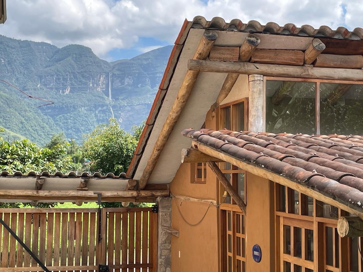 The exterior of the rustic house features a sloping roof with clay tiles and wooden beams. Large windows provide a view of the surrounding mountains, while a wooden fence encloses the property, enhancing the connection to nature.