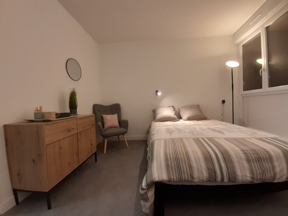 A comfortable sleeping area is furnished with a spacious bed dressed in neutral tones and a decorative striped blanket. A modern dresser stands nearby, accompanied by a cozy chair and a floor lamp. Natural light enters through a large window, enhancing the bright ambiance.