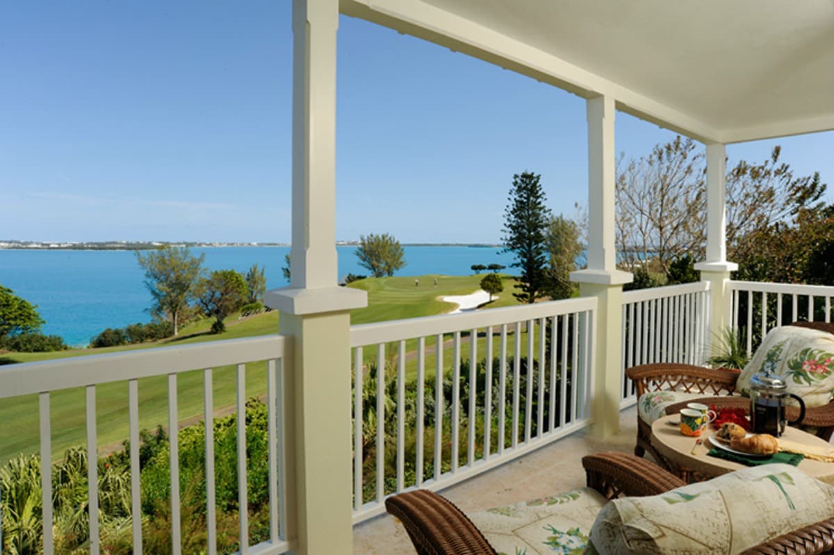 A covered veranda offers stunning views of the turquoise sea and lush greenery. Comfortable outdoor seating is arranged on the patio, complemented by a small table displaying a beverage and snacks, inviting relaxation while enjoying the scenery.