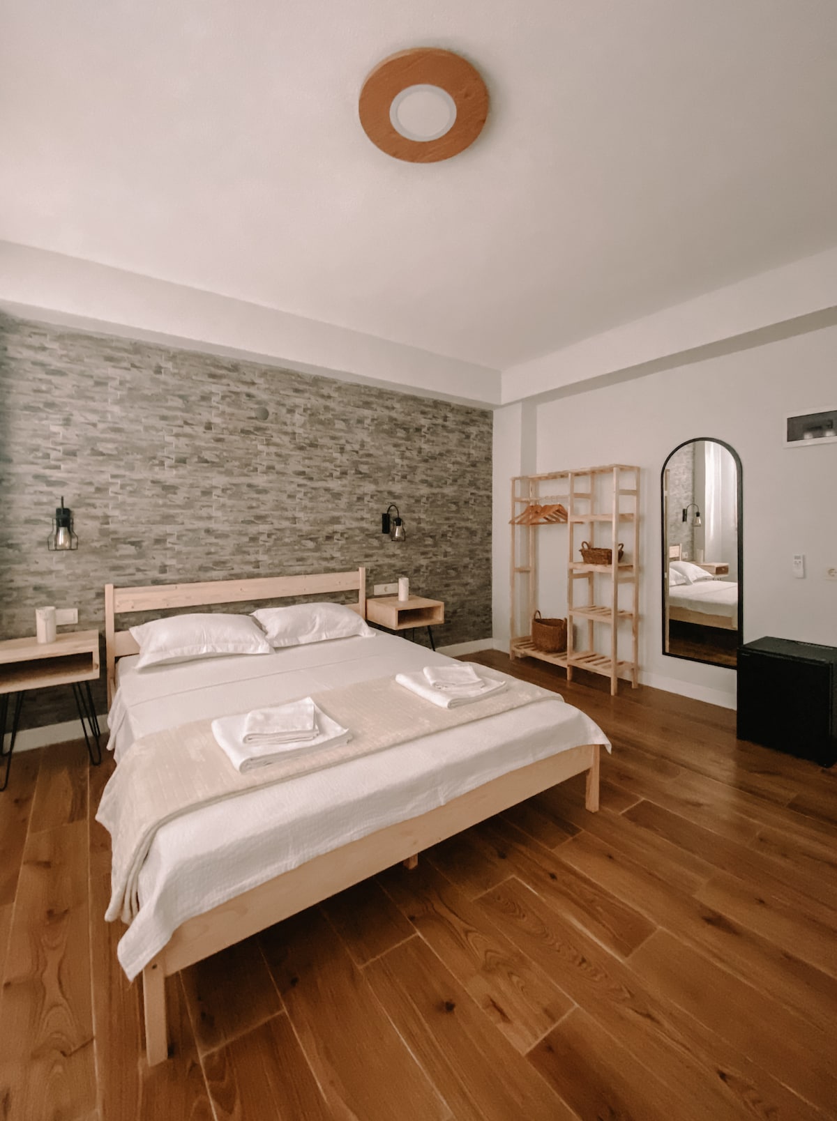 A spacious room features a large double bed with neatly arranged linens. Soft lighting illuminates the stone accent wall, while wooden furniture, including nightstands and a wardrobe, enhances the natural feel. A full-length mirror is positioned across from the bed, reflecting the minimalist décor.