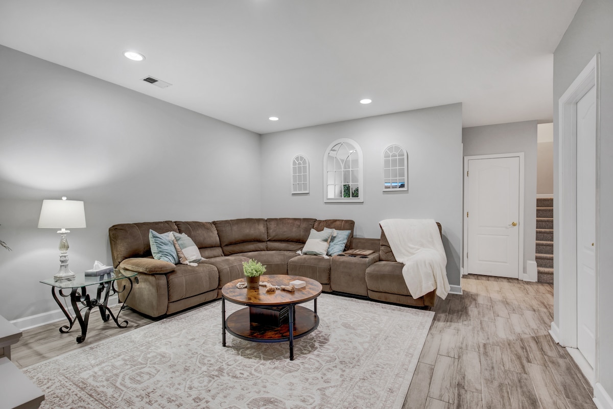The living area features a spacious sectional sofa adorned with soft pillows. A round coffee table sits centrally on a patterned rug, complemented by a lamp on a metal side table. Light emanates from recessed ceiling lights, illuminating the neutral tones of the walls.