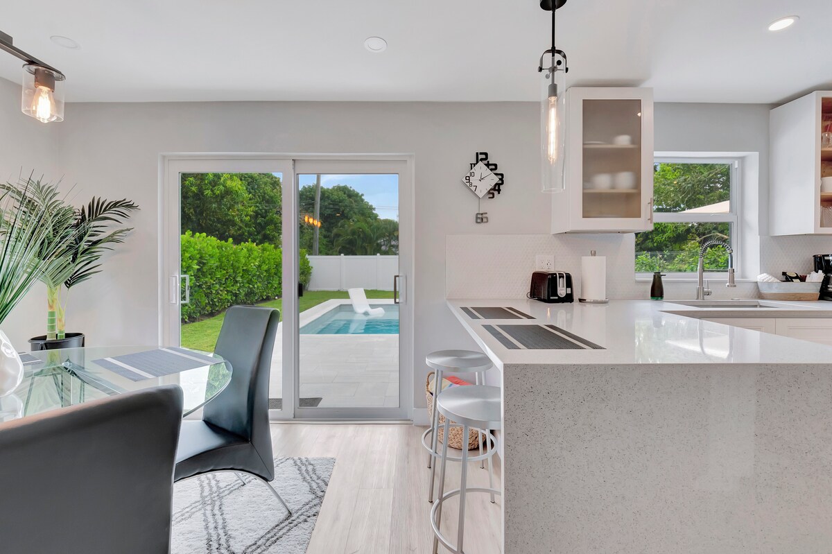 A bright kitchen area is seen, featuring a large island with sleek countertops and modern bar stools. Glass doors open to the backyard pool, offering a view of the outdoor space. Natural light fills the room through ample windows, accentuating a contemporary design.