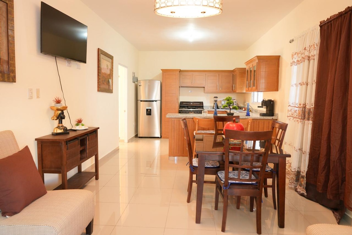 An open-plan kitchen and dining area features wooden cabinetry and stainless steel appliances, including a refrigerator and stove. A wooden dining table is surrounded by chairs, while a cozy seating area includes a modern armchair. Natural light brightens the space through large windows.