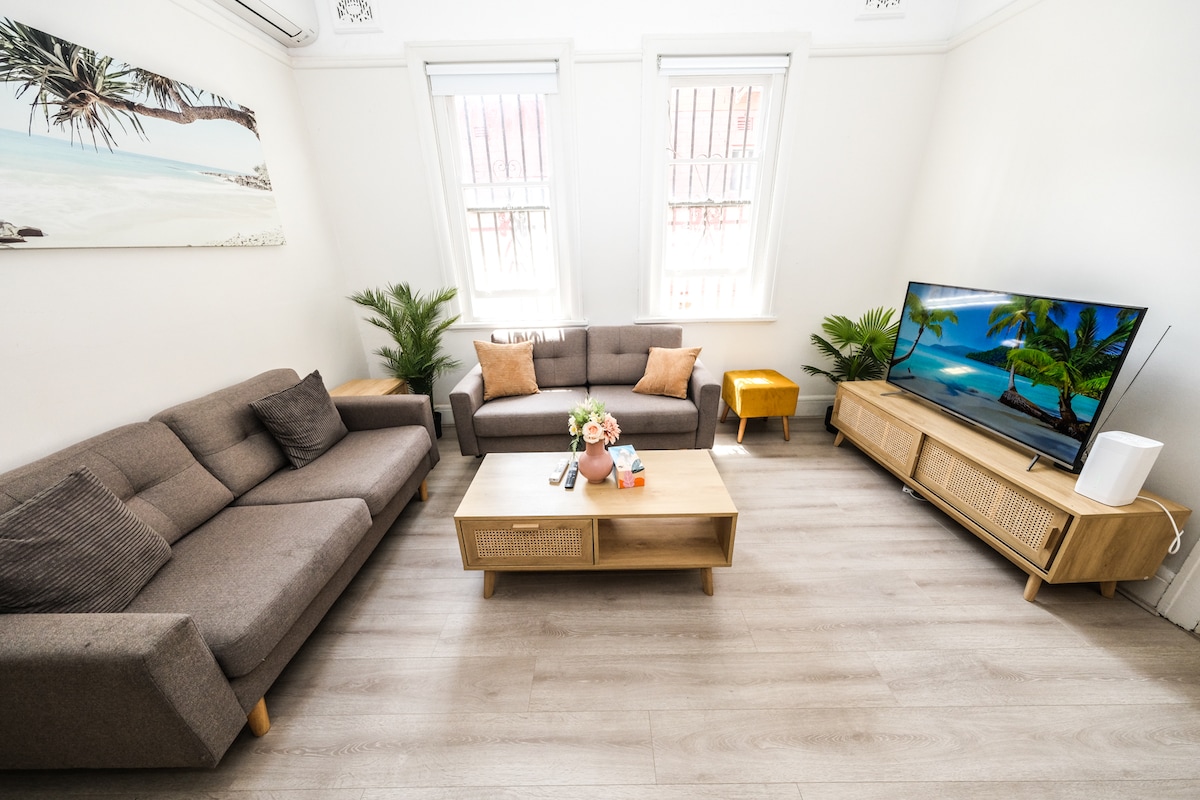 A thoughtfully arranged living area showcases two comfortable sofas paired with a wooden coffee table. A large flat-screen TV is positioned on a media console. Light fills the space through two windows, with decorative plants adding a touch of greenery.