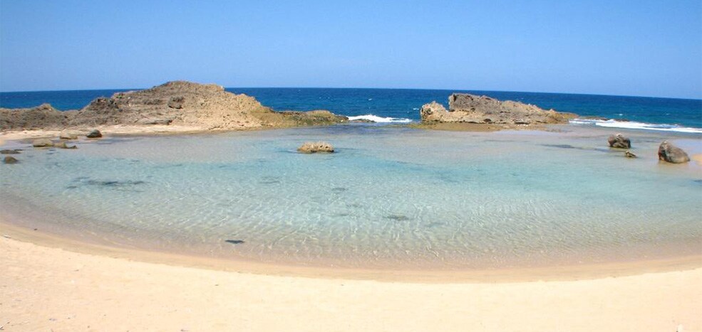 A serene beach scene features clear, shallow waters lapping against the sandy shore, with scattered rocks visible in the distance. The horizon stretches across a clear blue sky, creating a calm and inviting atmosphere for relaxation.