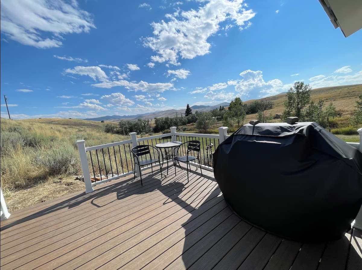 A spacious outdoor deck showcases a grill under a protective cover and a small dining set with two chairs and a table. The surrounding landscape features rolling hills and a blue sky with scattered clouds, creating a tranquil outdoor space.