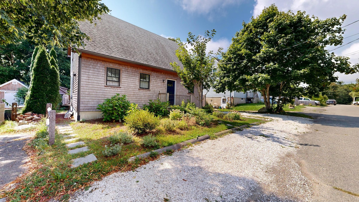 The exterior of a newly built two-story home is featured, set on a quiet residential street. Lush greenery surrounds the property, complemented by a gravel driveway. The front entrance is highlighted, while the charming architectural details of the house blend with the tranquil setting.