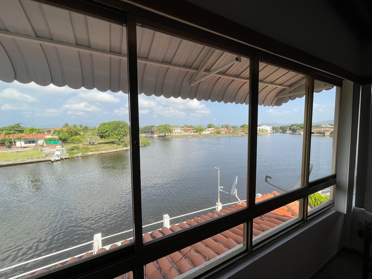 The view from the window showcases a calm canal bordered by greenery and residential buildings. A small boat can be seen docked nearby, with light reflecting off the water surface, creating a serene outdoor scene.