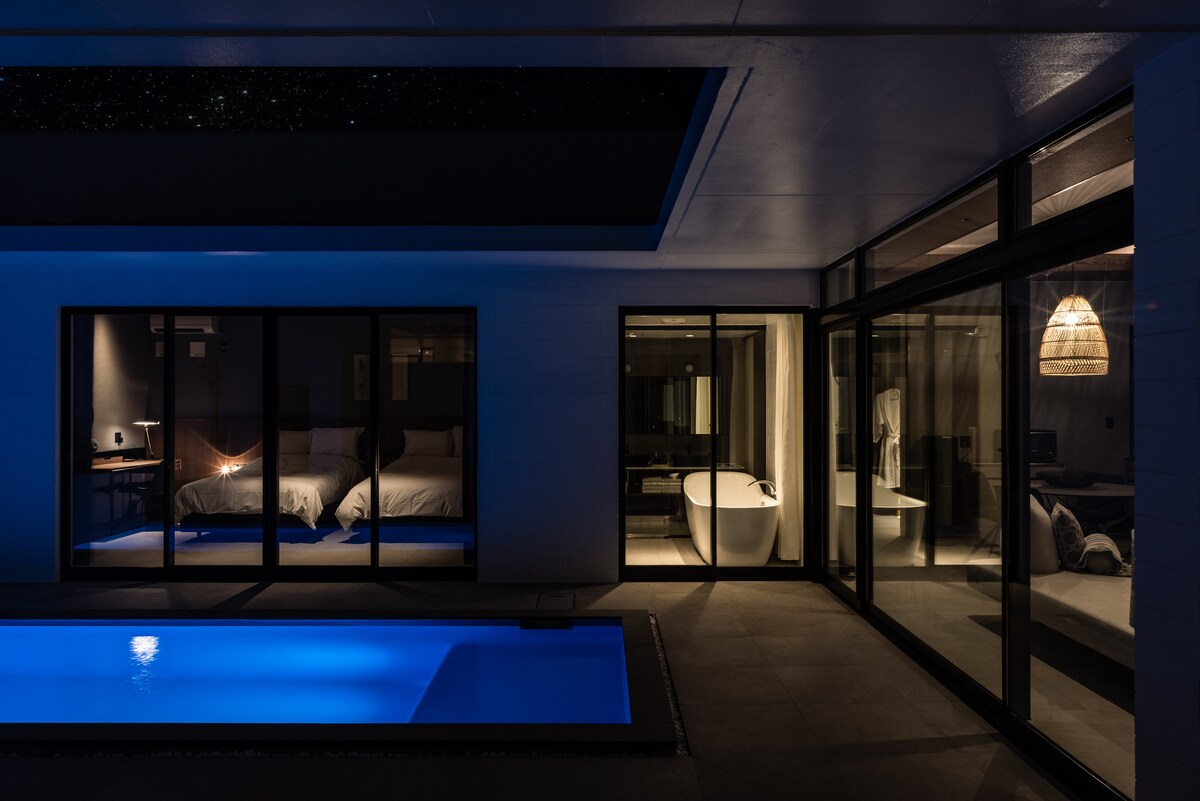 A private pool is illuminated in blue light, surrounded by a clean, modern design. Large glass doors offer views into a spacious bedroom with soft bedding and a designer bathtub. The ceiling features a starry motif, enhancing the serene ambiance of the villa.