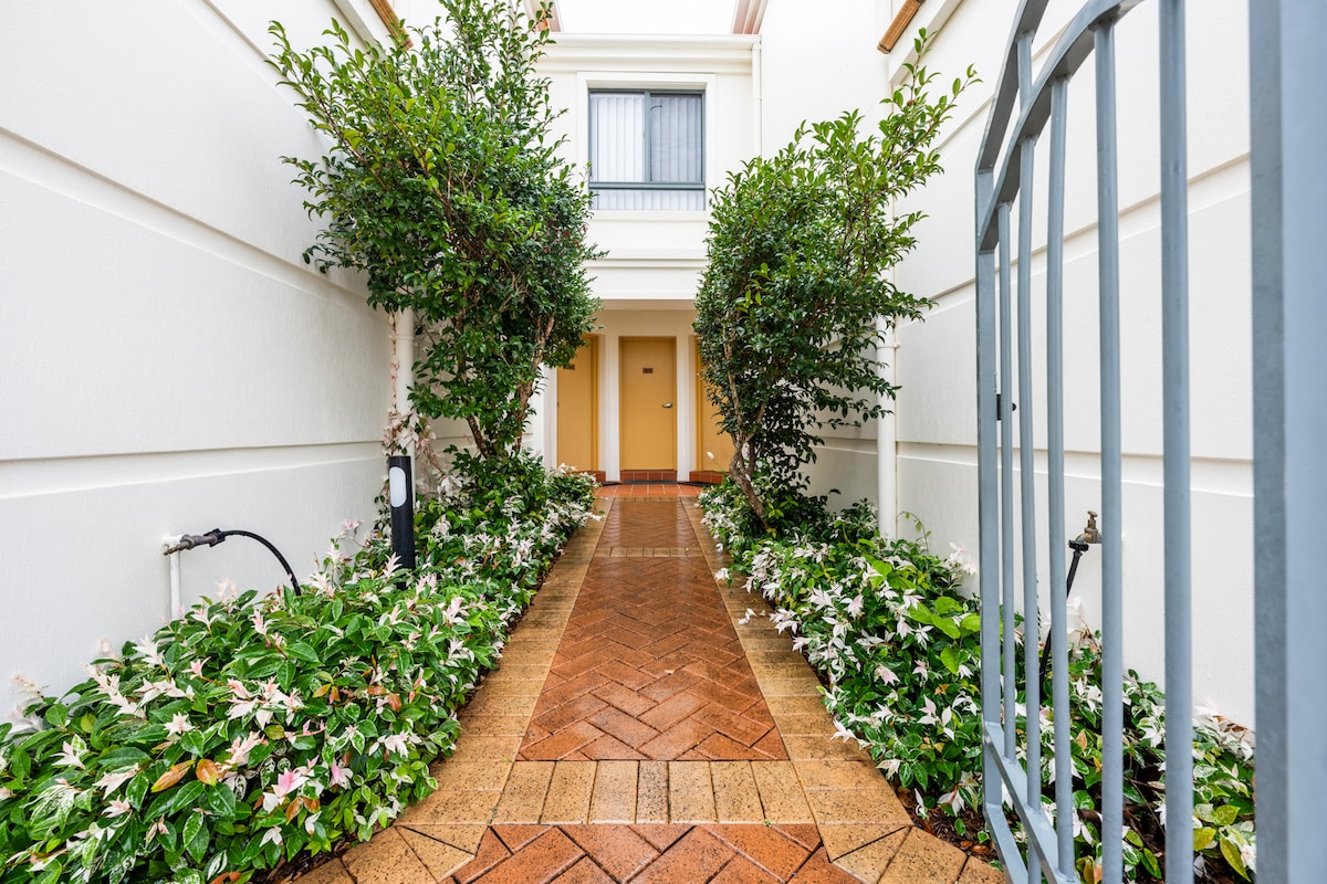 A landscaped pathway leads to the entrance of the townhouse, bordered by lush greenery and flowering plants. The walkway is paved with warm, earthy tiles, and the entrance door is framed by the building’s white exterior and a window above.