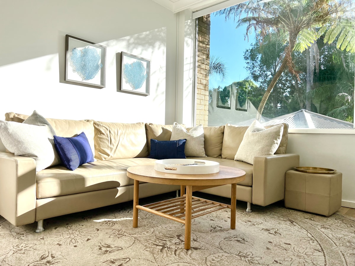 A spacious living area is illuminated by natural light. A light-colored sectional sofa is adorned with decorative pillows. A round wooden coffee table is positioned in front, with a small ottoman nearby. Large windows provide views of greenery outside.
