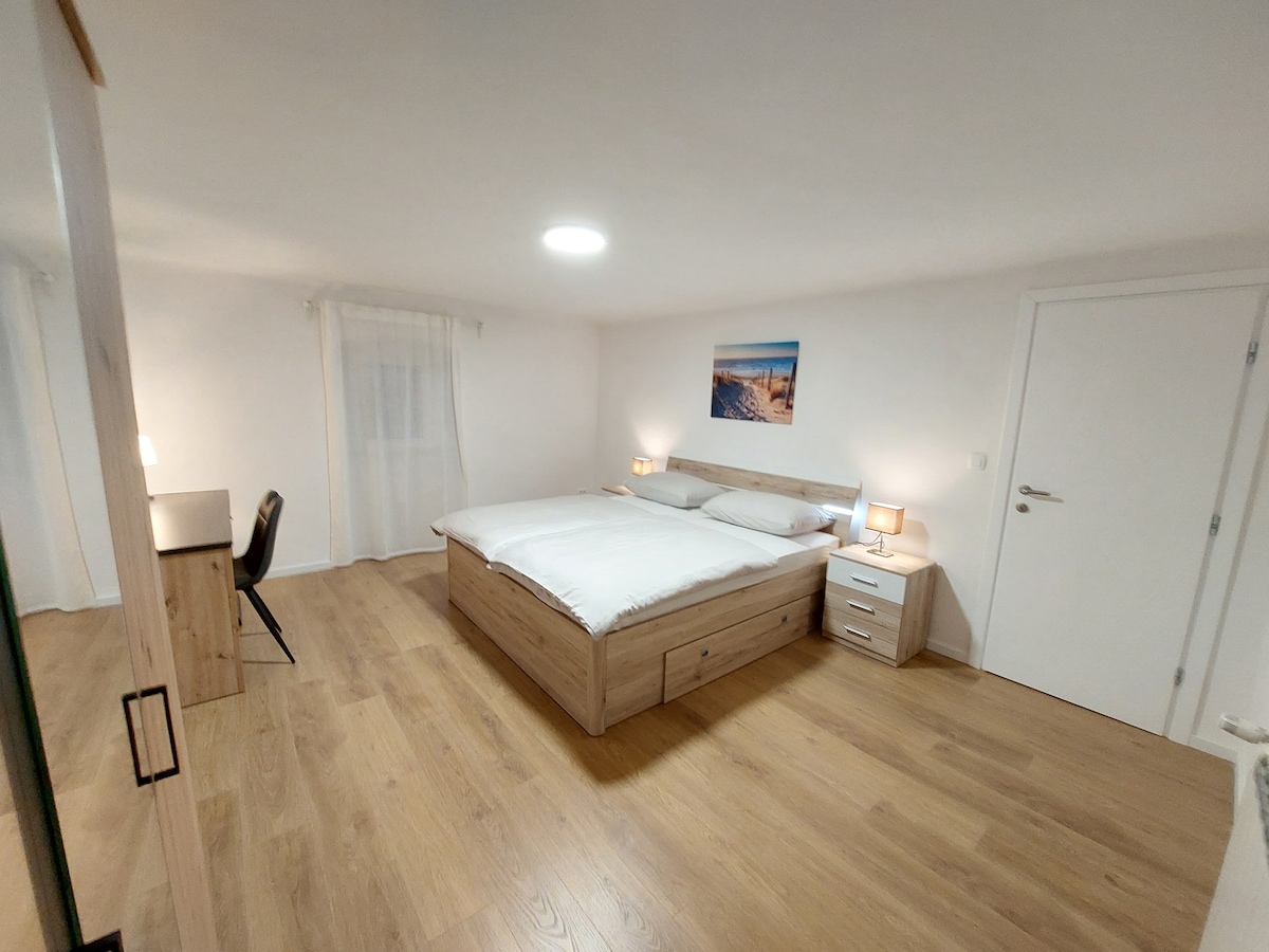 A spacious bedroom features a large bed with a minimalistic wooden frame and a light-colored bedspread. A simple desk and chair are situated near a window, complemented by a piece of wall art above the bed. Soft lighting is provided by bedside lamps.
