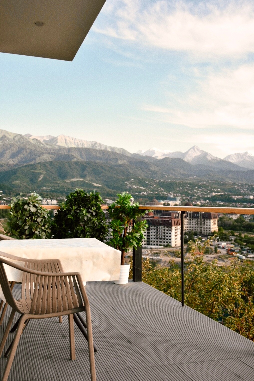 A furnished terrace is depicted, featuring a table and two chairs. Lush green plants border the area, enhancing the view of the mountainous landscape in the background. The sky is clear with soft clouds, creating a serene atmosphere.