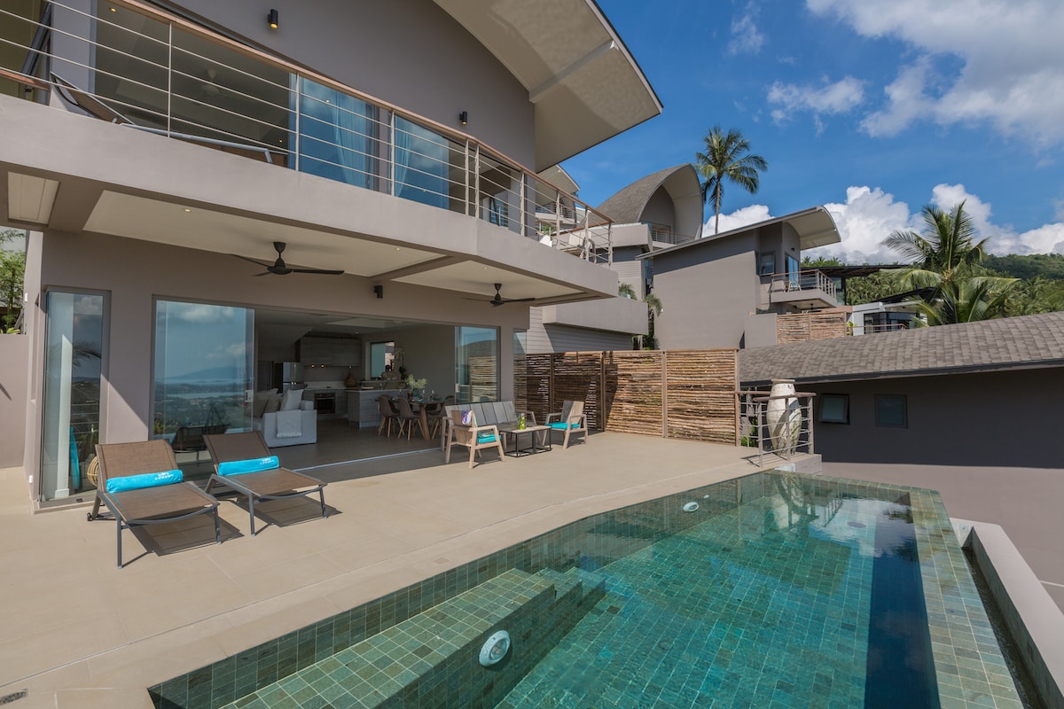 The outdoor area features a private swimming pool with a tiled surface that reflects the surroundings. Sun loungers are positioned nearby for relaxation. An open-plan space includes large glass doors that connect the indoor living area to the exterior, inviting ample natural light.