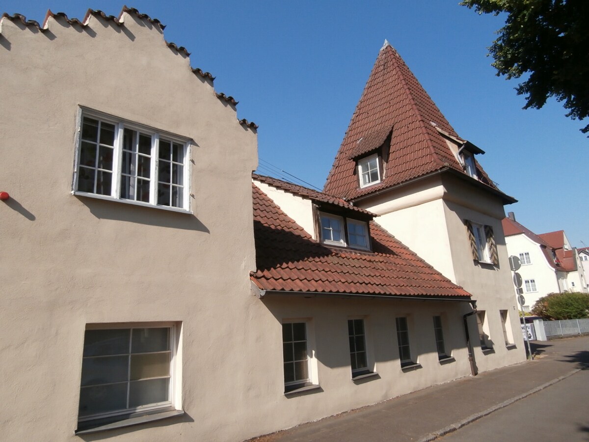 A two-part structure is displayed, with a tall turreted section on the right and a lower adjoining building on the left. The exterior features a light-colored facade with tiled roofs and multiple windows, showcasing a blend of architectural styles under clear blue skies.