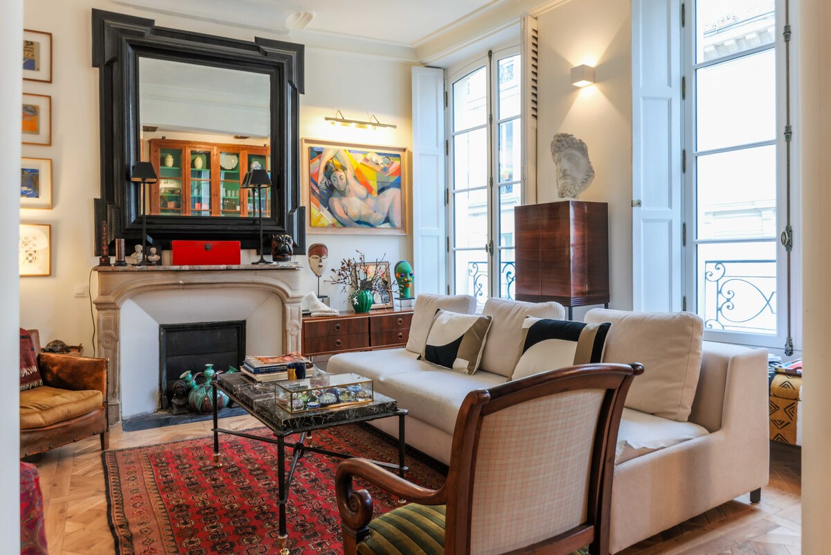 The inviting living area features a comfortable sofa with decorative cushions, positioned in front of a classic fireplace. Large windows allow natural light to fill the space, highlighting a vibrant area rug and eclectic artwork. A stylish coffee table adds a functional touch to the room.