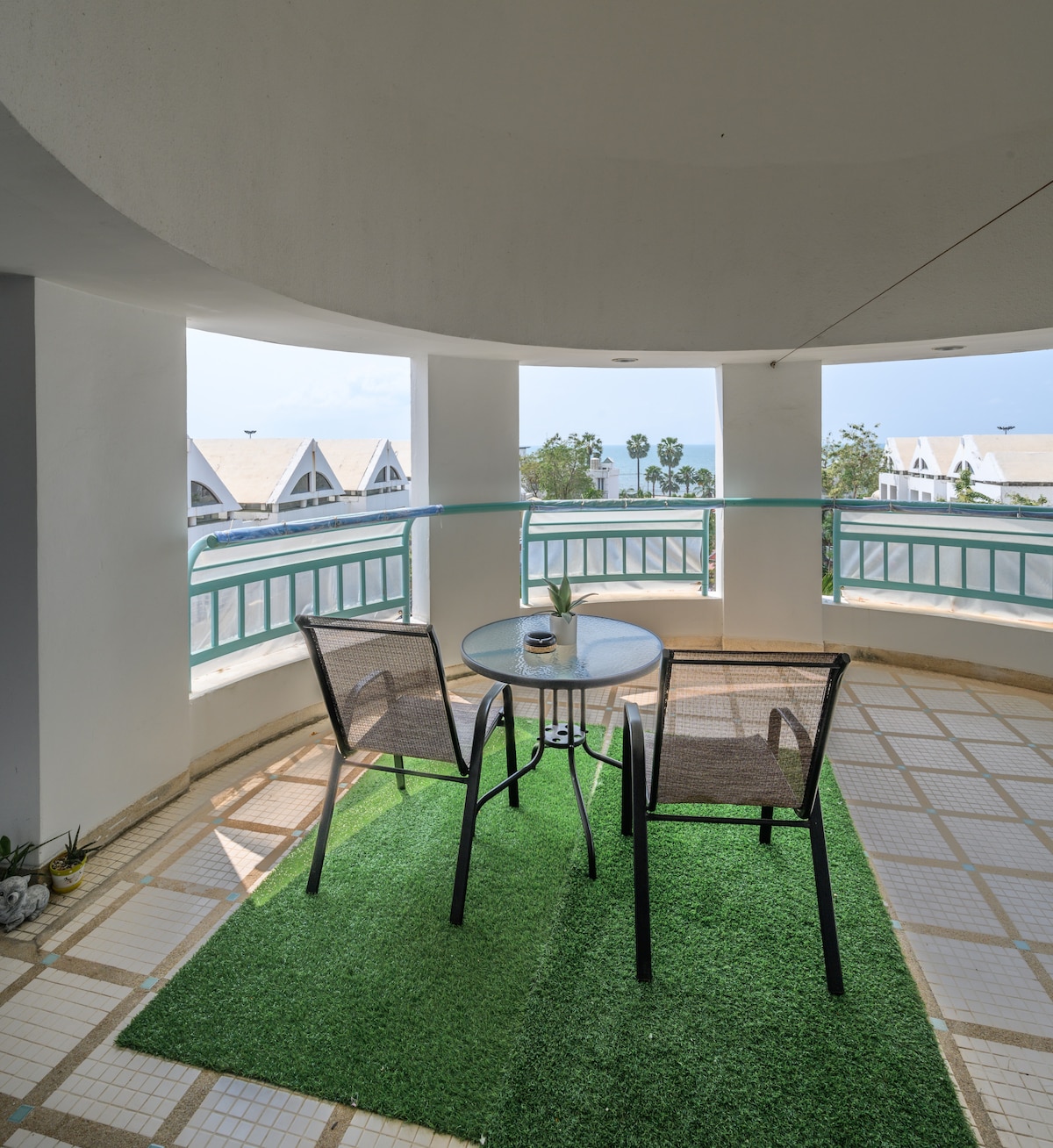 A circular balcony is shown, featuring a glass-top table surrounded by two black chairs. The area is adorned with artificial grass, offering a comfortable spot to enjoy a fresh breeze and views beyond the railing.
