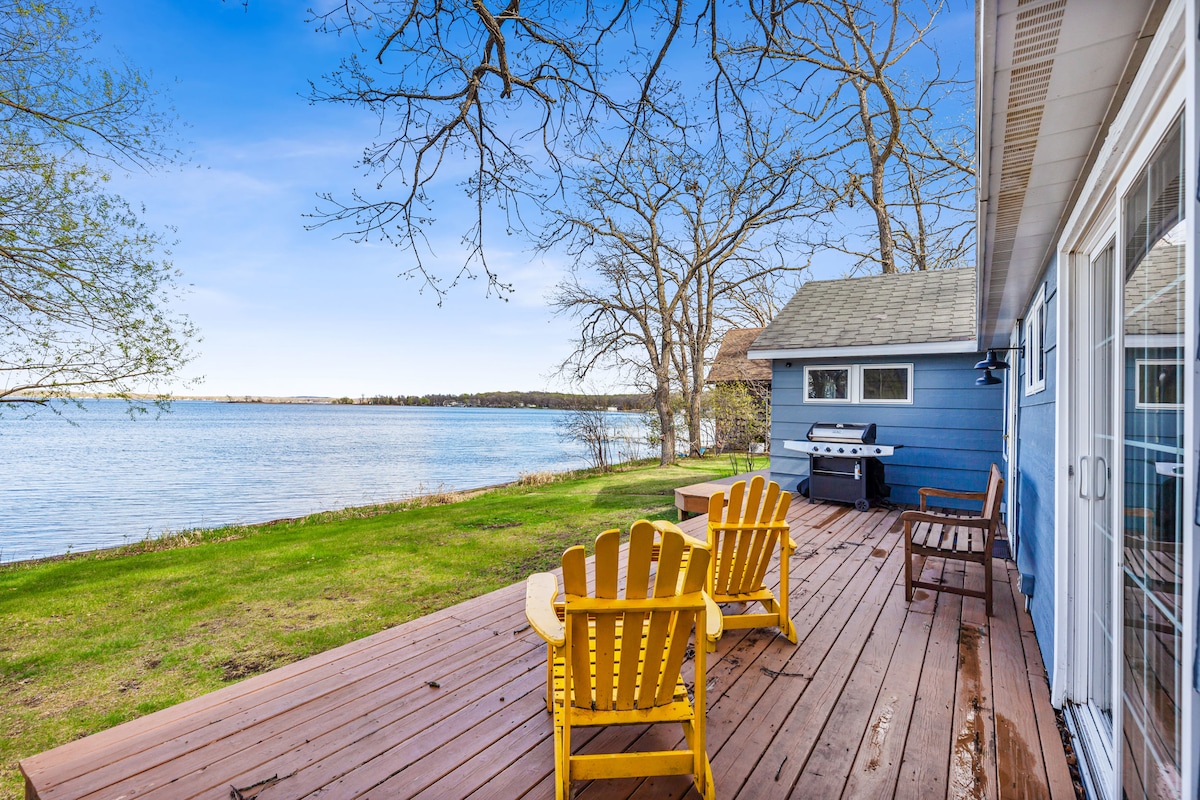 A deck is seen extending along the water's edge, featuring two yellow Adirondack chairs and a grill. A grassy area frames the wooden surface, while trees provide a natural backdrop. The tranquil lake reflects the clear sky, enhancing the outdoor space's charm.