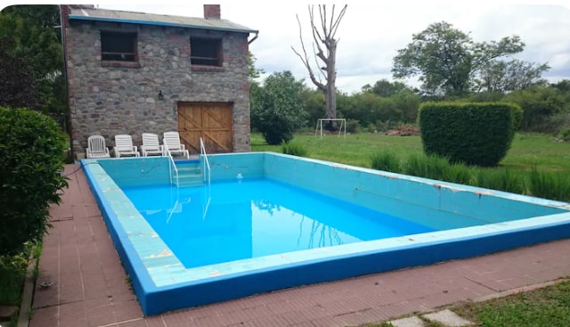 A large outdoor pool features clear blue water and is surrounded by a spacious patio. Several white lounge chairs are placed nearby, and lush greenery borders the area, providing a natural setting. A rustic stone building stands in the background.