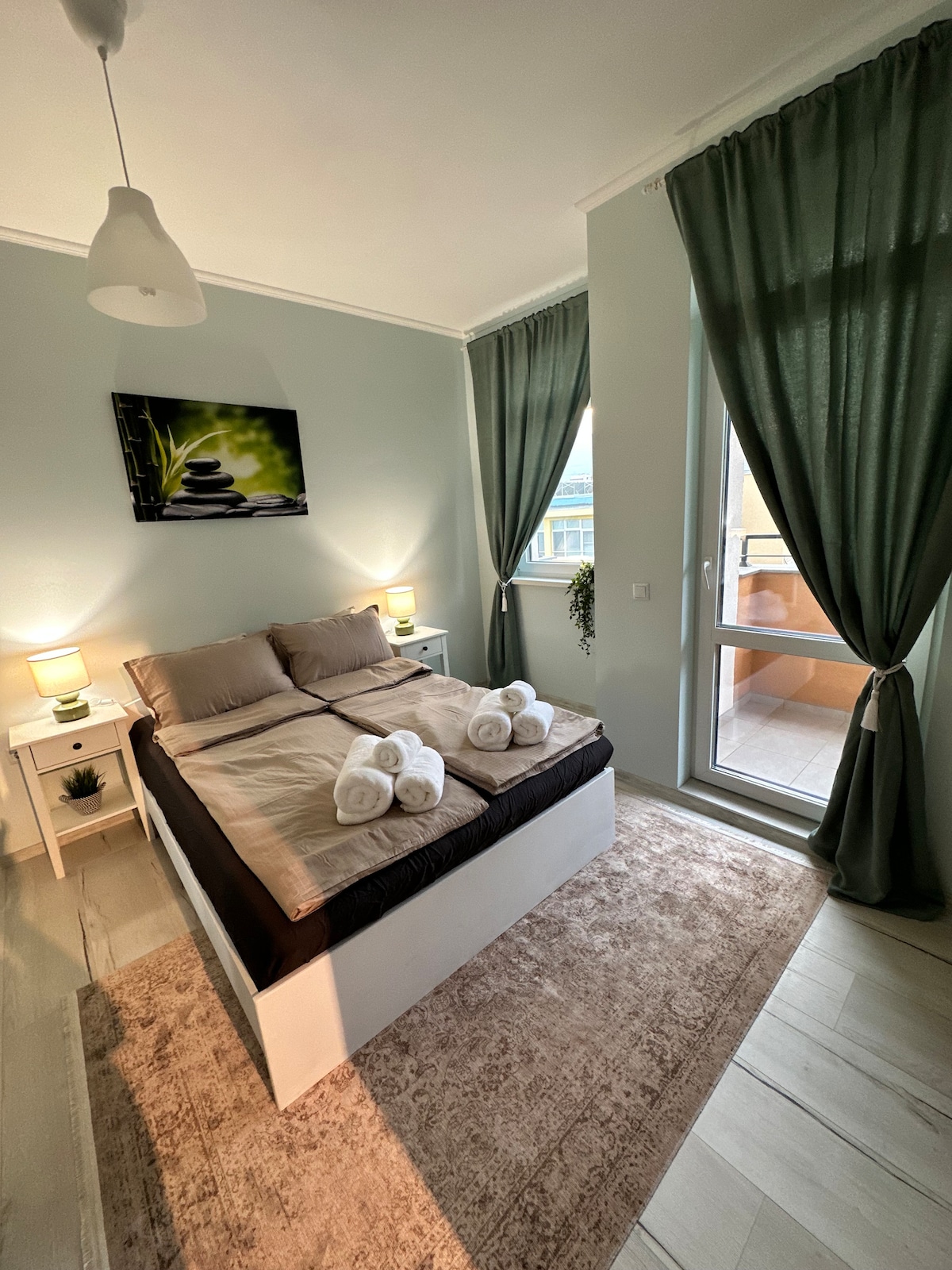 A spacious bedroom features a large bed dressed in neutral linens, accompanied by soft towels arranged neatly on top. Natural light enters from a window, complemented by green curtains and a decorative TV mounted on the wall. A small bedside table adds functionality.