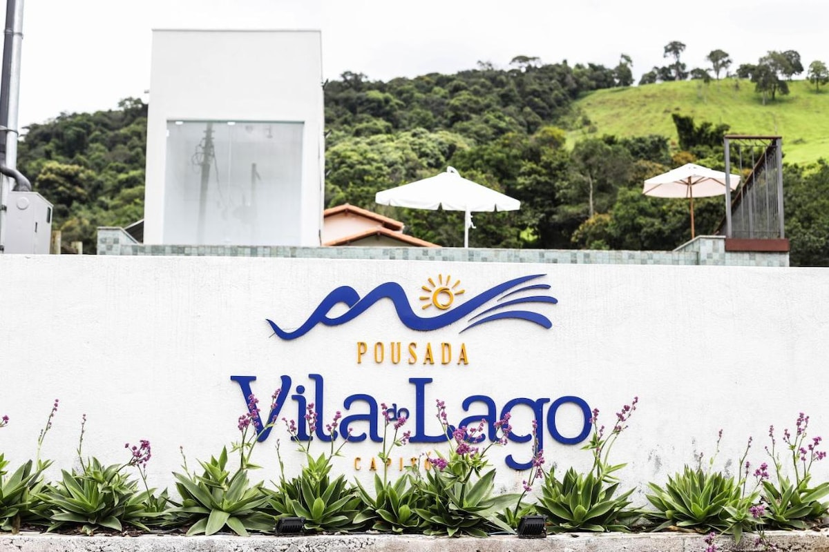 The entrance sign of Pousada Vila do Lago is prominently displayed against a backdrop of green hills. Surrounding the sign, decorative plants and flowers enhance the welcoming appeal, while umbrellas are seen in the distance, suggesting outdoor areas for relaxation.