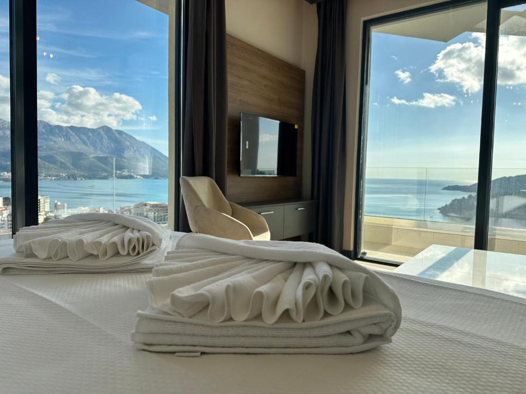 A spacious bedroom features a large window offering stunning views of the sea and mountains. Light-colored linens are neatly arranged on the bed, while a television is mounted on the wall. Soft natural light illuminates the area, complementing the cozy chair in the corner.